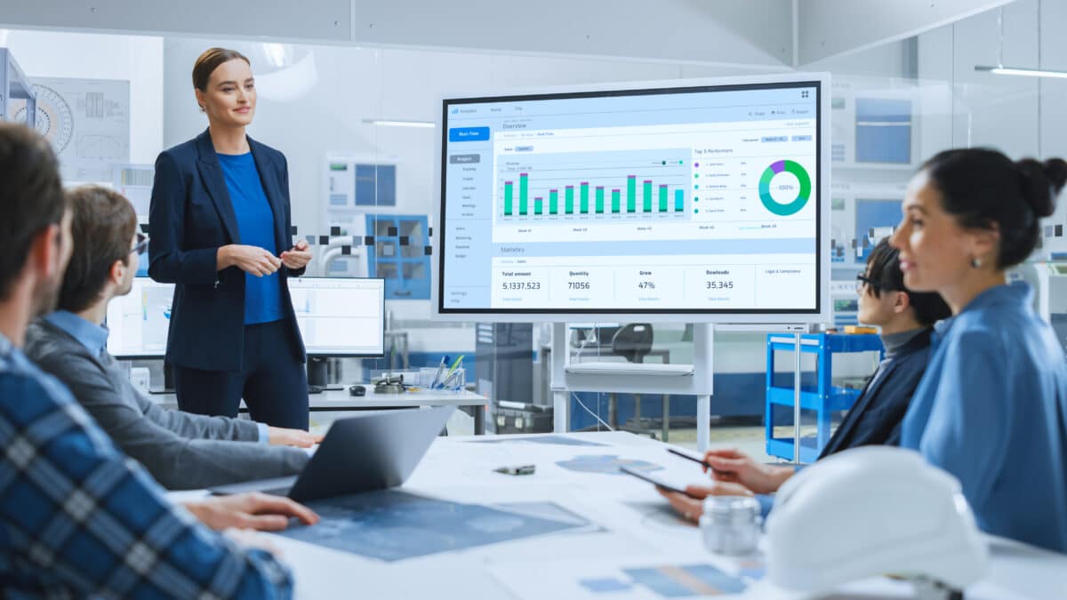 Modern Industrial Factory Meeting: Confident Female Engineer Uses Interactive Whiteboard, Makes Report To A Group Of Engineers, Managers Talks And Shows Statistics, Growth And Analysis Information